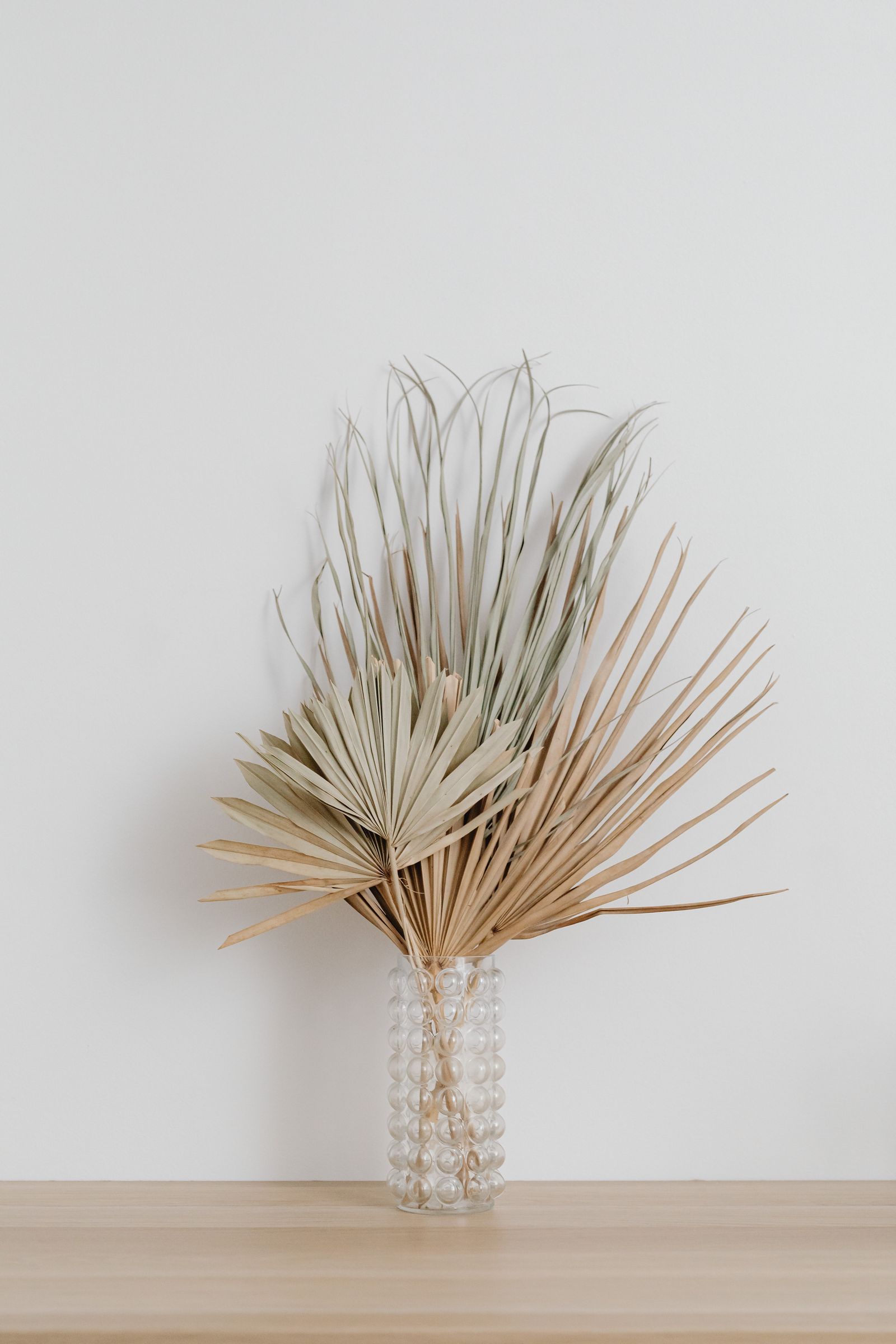 Dried palm leaves in a textured glass vase on a light wood shelf